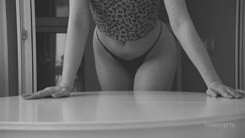 Slim woman in leopard print top and black bikini briefs leaning on a white table in a moody indoor setting.