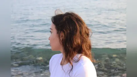 Side profile of a woman with wavy brown hair wearing a white shirt by the calm blue ocean at sunset.