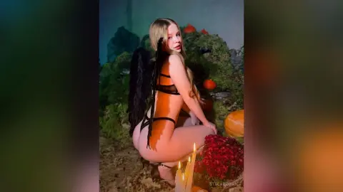 StelaBrown poses naked in a dark, mossy forest setting, wearing black strap lingerie and black wings while kneeling near lit candles.