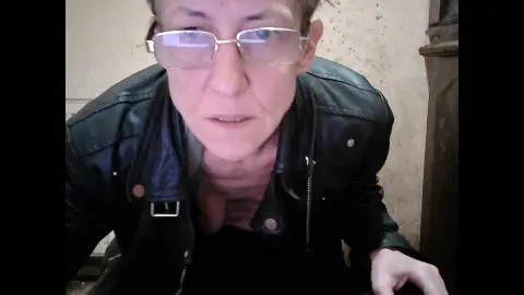 Close-up of a woman in a black leather jacket and glasses indoors with a neutral expression.