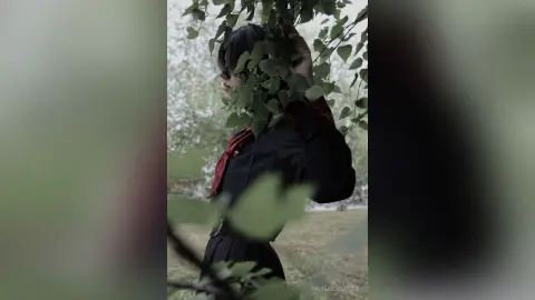 Moody portrait of a woman in a black outfit standing behind leafy green branches in a soft-focus garden.
