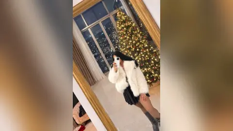 Young woman in a white faux fur coat posing in front of a decorated Christmas tree in a modern apartment.