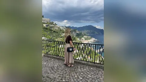 AmaFox — Blonde woman in a brown top and beige skirt overlooking the scenic Amalfi Coast cliffs under a cloudy sky.