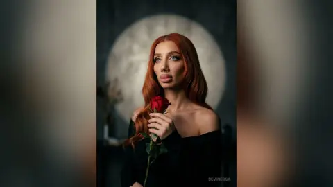DeviNessa holds a single red rose against her chest, wearing an off-the-shoulder black top with her long red hair flowing down.