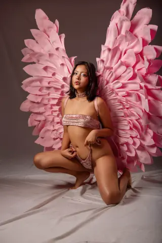 Young woman in a pink sequin bikini wearing large pink feathered angel wings in a dreamy studio setting.