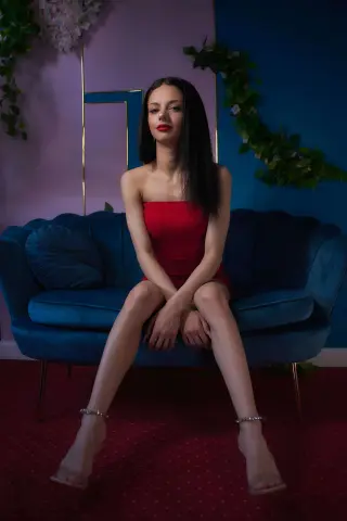 Elegant woman in a red cocktail dress sitting on a blue velvet sofa in a moody, stylishly lit room.