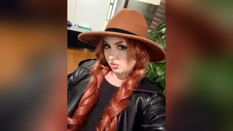 Redheaded woman in a black leather jacket and wide-brimmed brown hat posing with a moody aesthetic.