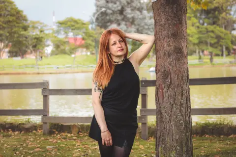 Redheaded woman in a black sleeveless dress leaning against a tree by a peaceful lake in a park.