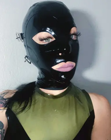 Woman in a black shiny latex mask and olive green bodysuit posing against a plain wall.