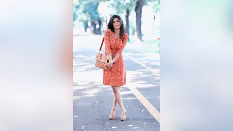 Stylish woman in a red polka dot midi dress and tan heels standing on a sunny park path.