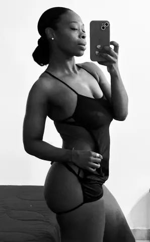 Black woman in black bodysuit posing for a moody indoor selfie against a white wall.