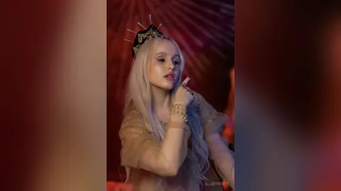 EllieFrosty poses with long silver hair wearing a gold crown and a sheer tan mesh top.