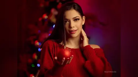 Smiling woman in a red sweater holding a Christmas ornament in front of a festive holiday tree background.