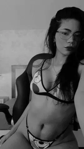 Young woman with glasses in a patterned bikini relaxing in a bedroom, black and white moody aesthetic.