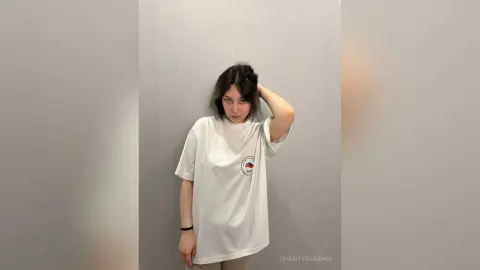 Young woman in a white oversized graphic t-shirt posing against a minimalist grey wall with a relaxed mood.