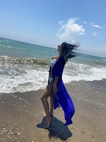 Issadorra stands on a sandy beach wearing a long, sheer blue robe, letting her dark hair blow in the wind by the ocean waves.