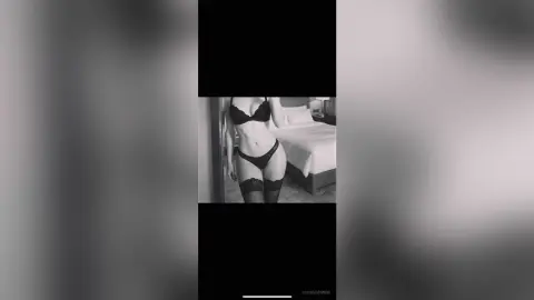 AlmaShannon poses in black lace lingerie, wearing a bra and panties with lace garters, standing next to a bed in a bedroom setting.