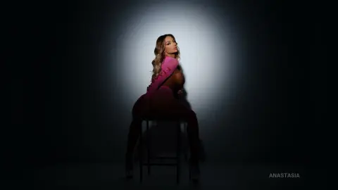 Anastasia poses naked on a black stool under a bright spotlight, showing her bare chest and hips.