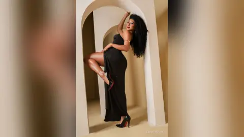 Elegant woman in a sleek black evening gown posing confidently against a minimalist beige arched backdrop.