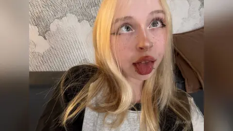 Blonde woman with facial piercings sticking out her tongue in a cozy indoor setting.