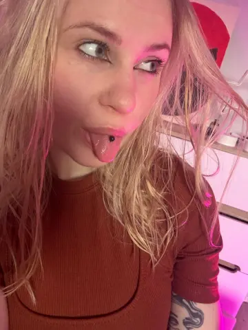 Blonde woman in a brown ribbed top making a playful expression in soft pink lighting.