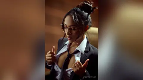 ValerySinclair wears glasses and an open white shirt with a black tie, looking down while posing in a dark room.