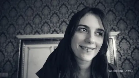 Smiling young woman with long dark hair and facial piercings in a vintage room with patterned wallpaper.