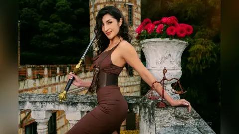 VeronicaValdez poses outdoors in a brown sleeveless dress, leaning against a stone balcony next to a white flower vase.