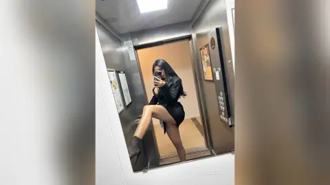 NicoleBella poses in a mirror reflection wearing a black dress and sheer black stockings while standing in an elevator.