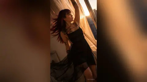 Tattooed woman in a black slip dress posing by a window during a warm, golden hour sunset.