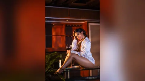 Elegant woman in a white silk shirt sitting in a moody, dimly lit luxury lounge holding a glass of red wine.