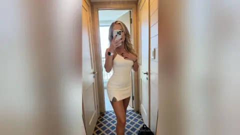 Young woman in a white mini dress taking a mirror selfie in a bright, modern hallway.