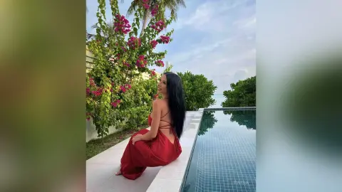 Smiling woman in a red backless dress sitting by a luxury poolside garden with pink bougainvillea flowers.