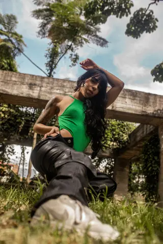 Young woman in a green ribbed tank top and black trousers posing outdoors in a sunny garden setting.
