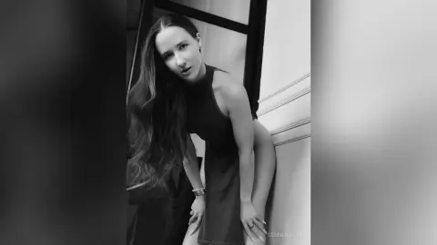 Elegant woman in a black sleeveless dress posing in a moody black and white indoor setting.