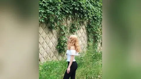Blonde woman in a white off-the-shoulder top and black pants standing by a lush green ivy wall.