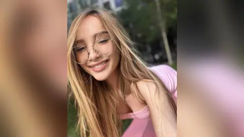 Smiling young woman with long blonde hair and gold glasses wearing a pink top in a sunny outdoor setting.