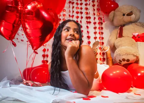 Smiling young woman in white loungewear lying amidst red heart balloons and a teddy bear for Valentine's Day.