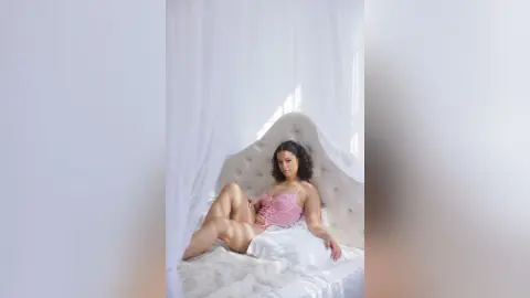 Curvy woman in pink lace lingerie relaxing on a white bed in a bright, airy bedroom.