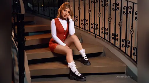 Young woman in a red vest and white long-sleeve top sitting on indoor stairs with a moody, aesthetic vibe.