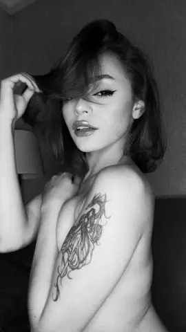 Black and white portrait of a young woman with long hair and a large chest tattoo in a moody indoor setting.
