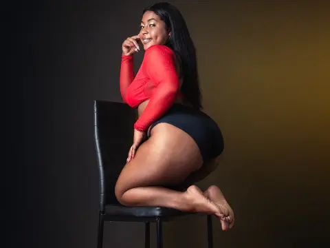 Smiling woman in a red long-sleeve crop top and black shorts sitting on a chair against a dark studio background.