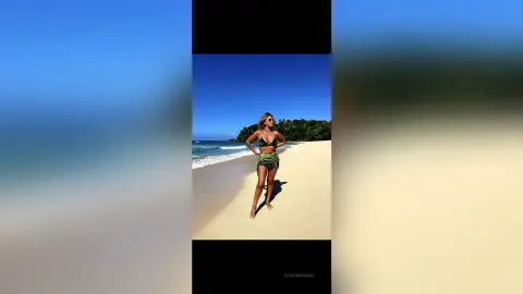 Stylish woman in a green bikini and shorts standing on a sunny tropical beach under a clear blue sky.