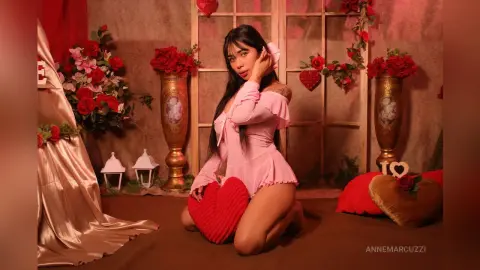 Woman in a pink ruffled bodysuit kneeling among red roses and heart decor in a romantic, warm setting.