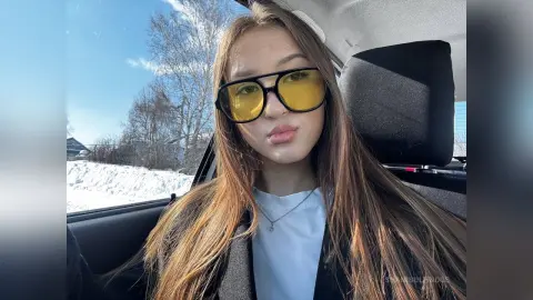 Young woman with long hair wearing yellow sunglasses and a white shirt sitting in a car on a sunny day.