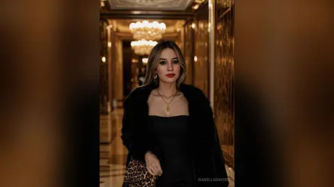 Elegant woman in a black dress and faux fur coat posing in a luxury hotel hallway with warm lighting.