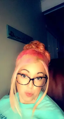 Woman with blonde and pink hair wearing glasses and a turquoise t-shirt in a dimly lit room.