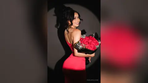 Smiling woman in a red evening gown holding a large bouquet of red roses against a dark studio background.