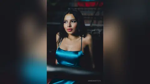 Elegant woman in a teal satin slip dress posing with a moody, cinematic look in a dimly lit setting.