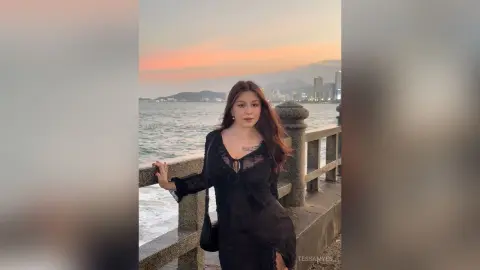 Young woman in a black lace dress standing by a seaside railing during a dreamy sunset.